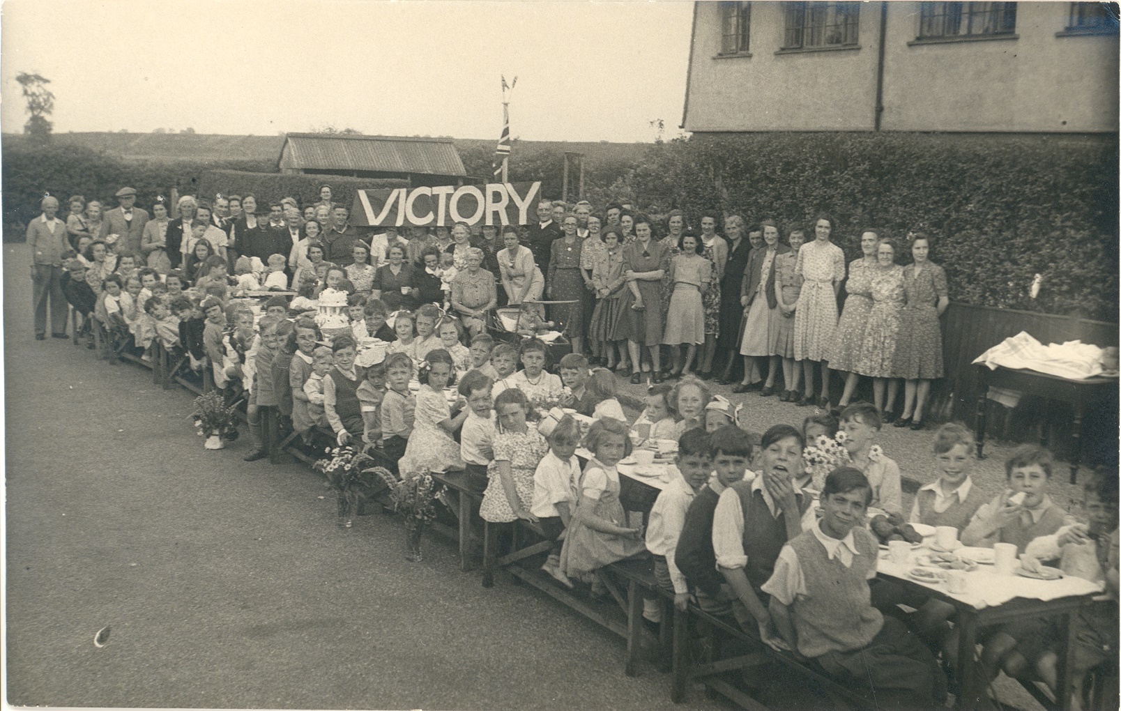 Victory celebrations in Florence Road, Walton, showing a long table with children sitting at it and adults in the background holding a 'VICTORY' banner.